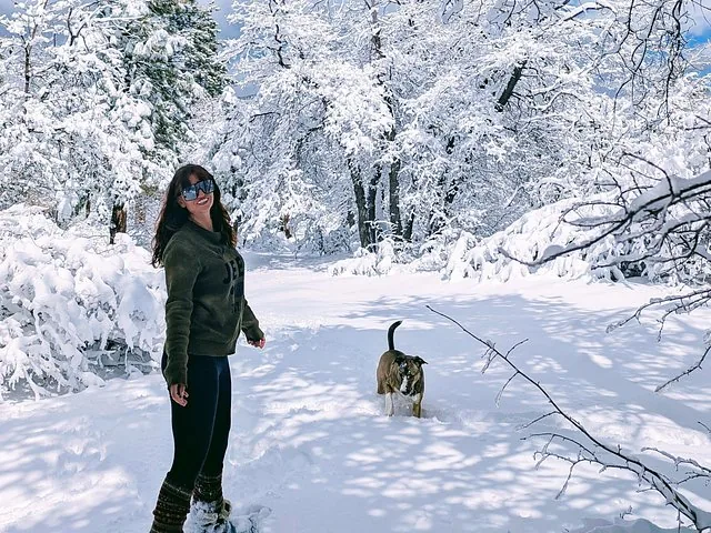 A woman stands in a snowy forest clearing with a dog nearby.